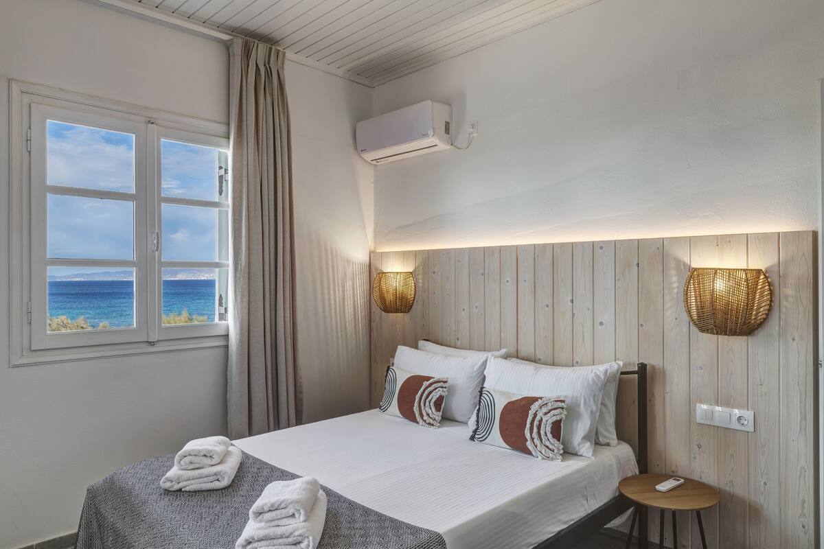 A hotel room with a bed, a view of the sea, an air conditioning unit, and a small table with a lamp.