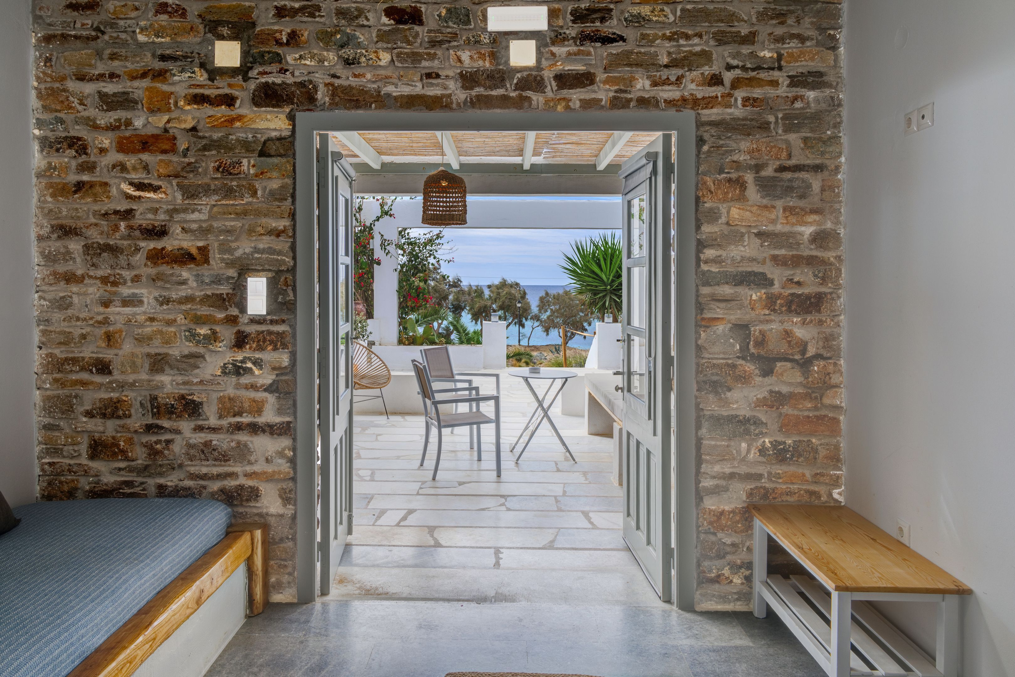 Una stanza con parete in pietra, un letto, una panca e vista sul mare attraverso una porta aperta.