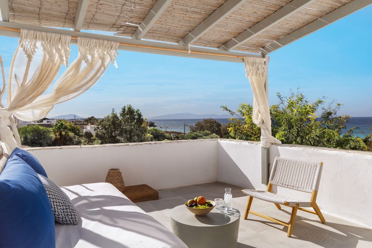 A balcony with a view of the sea, a wicker chair, a small table with fruit and a glass, and a sofa with cushions.