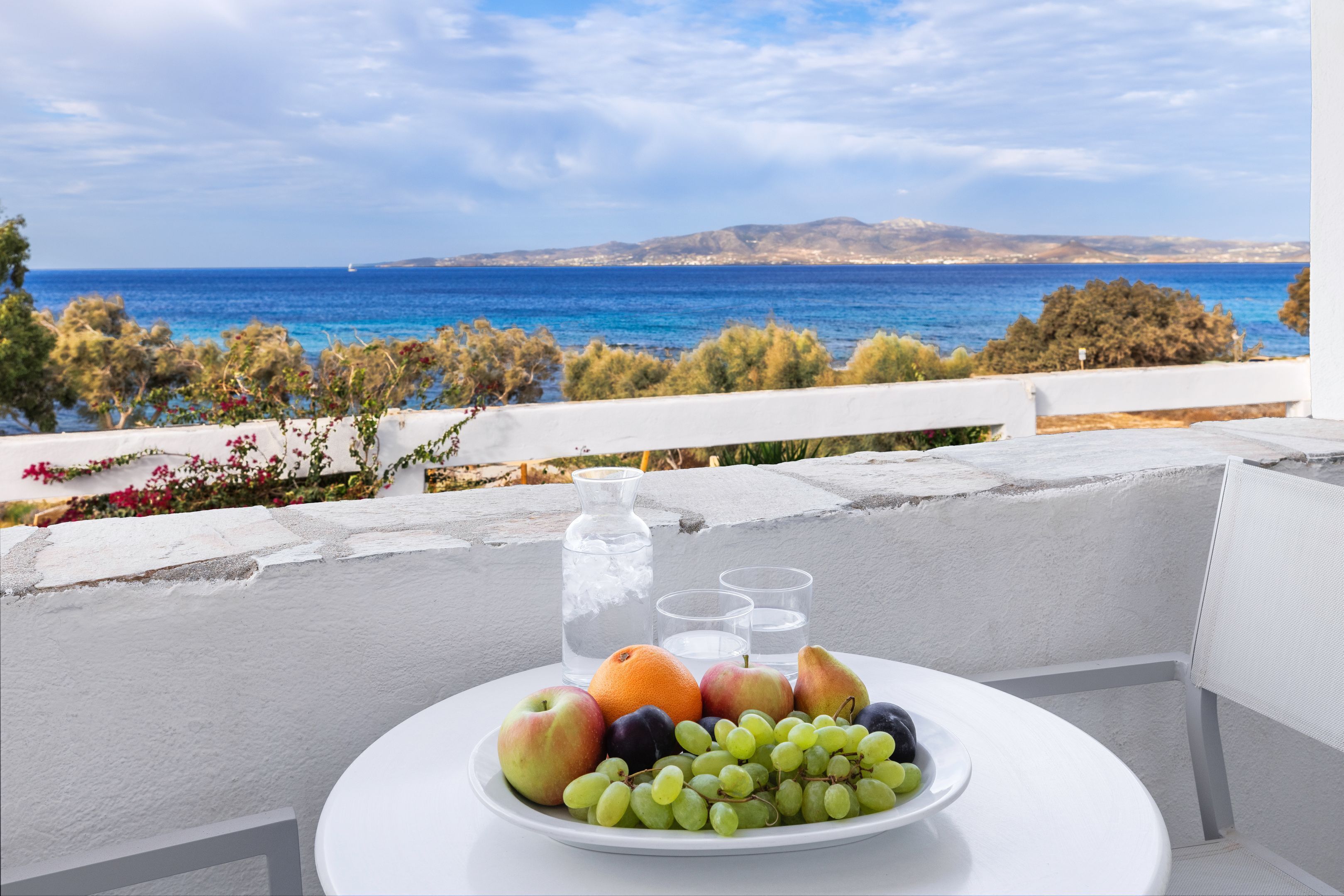 A table with a fruit platter, a water pitcher, and glasses on a balcony overlooking the sea.