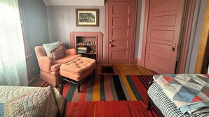Ein gemütliches Zimmer mit einem rosa Sessel, einem Bücherregal, einem farbenfrohen Streifen Teppich und zwei rosafarbenen Türen.