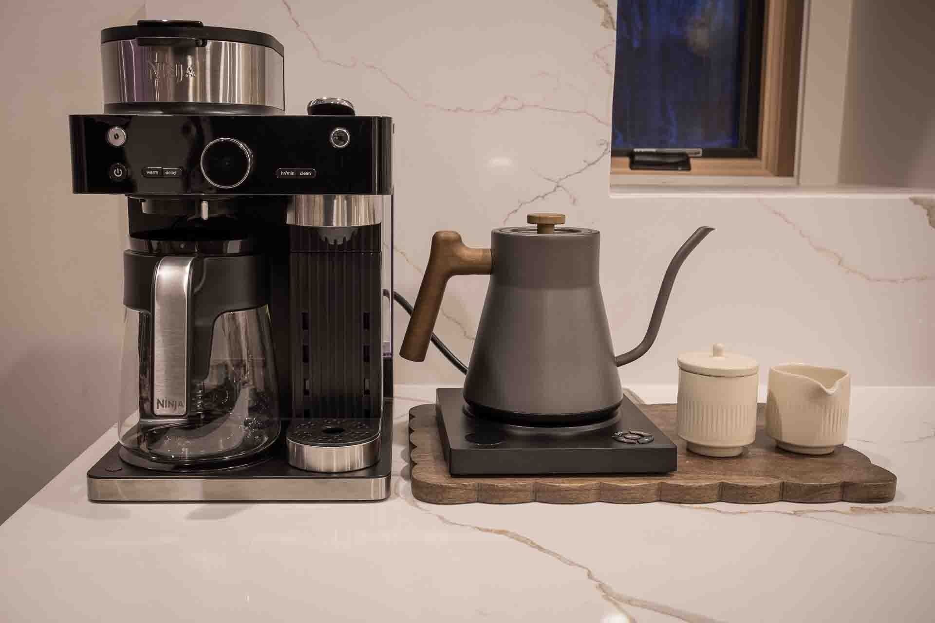 A coffee maker with a water reservoir, a kettle, and two white cups on a wooden tray.