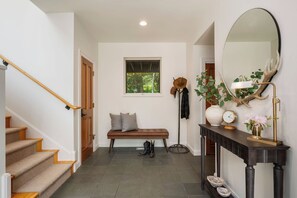 A staircase with wooden steps and a handrail, a bench with cushions, a wall-mounted mirror, a clock, and a vase with flowers.