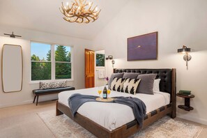 A bedroom with a large bed, a bench, a chandelier, a painting, and a view of trees through the window.