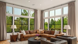 A spacious living room with a sectional sofa, a round coffee table, and a radiator. Large windows provide a view of greenery outside.