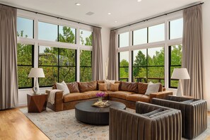 A spacious living room with a sectional sofa, a round coffee table, and a radiator. Large windows provide a view of greenery outside.