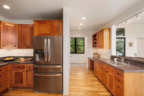 A modern kitchen with wooden cabinets, a stainless steel refrigerator, granite countertops, and a window with a view of greenery.