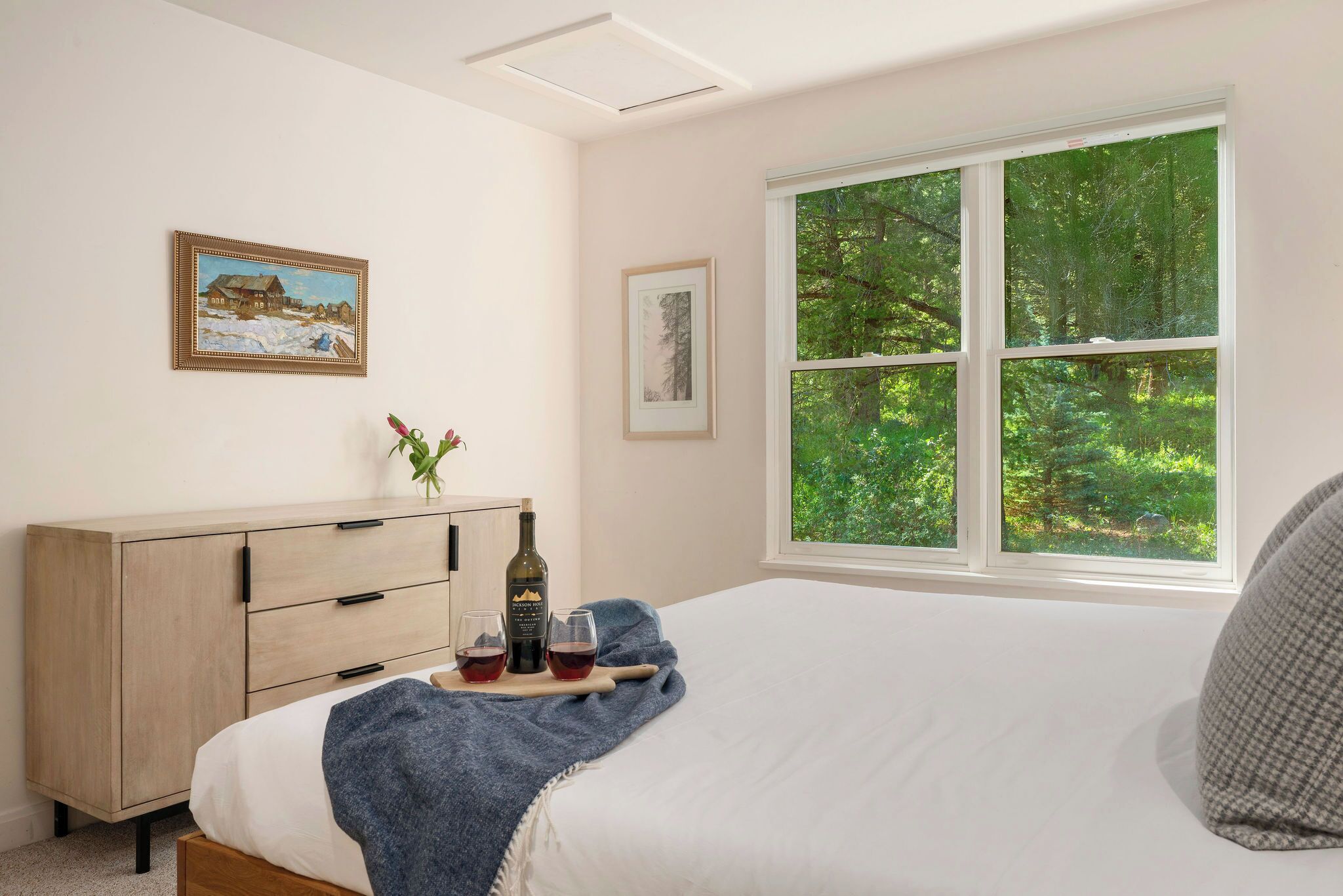 Une chambre à coucher comprenant un lit, une commode, une fenêtre donnant sur une forêt et deux tableaux encadrés accrochés au mur.