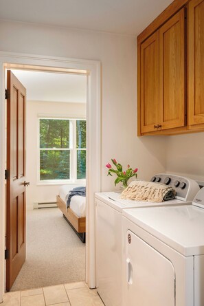 A laundry room with a washer and dryer, wooden cabinets, a bed with a view to a forest, and a door leading to another room.