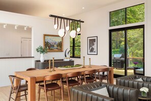 A modern dining area with a wooden table and chairs, a hanging light fixture, and a view of greenery through large windows.