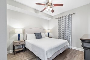 A bedroom with a bed, two bedside tables with lamps, a ceiling fan, and a patterned curtain. - San Carlos 1905 (Gulf Shores)