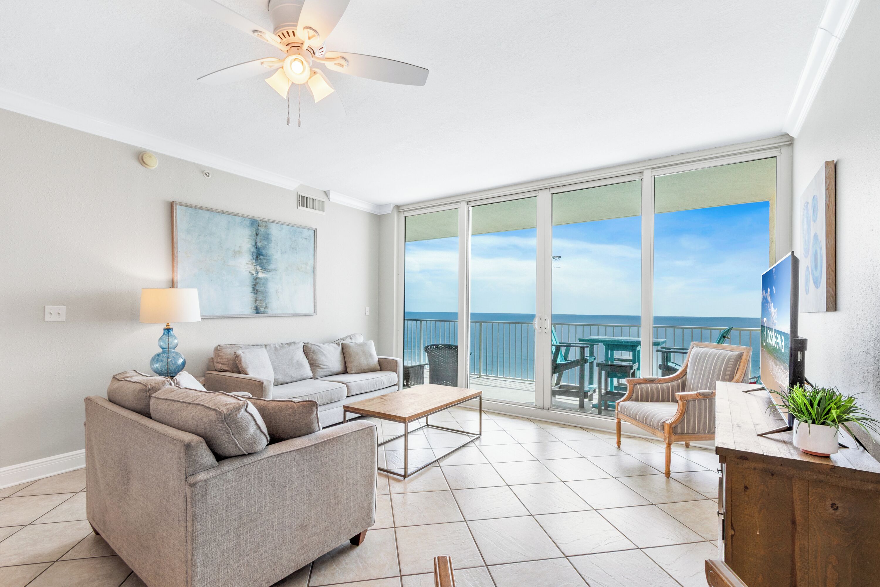 A living room with a sofa, coffee table, and a TV. There is a balcony with a view of the ocean.