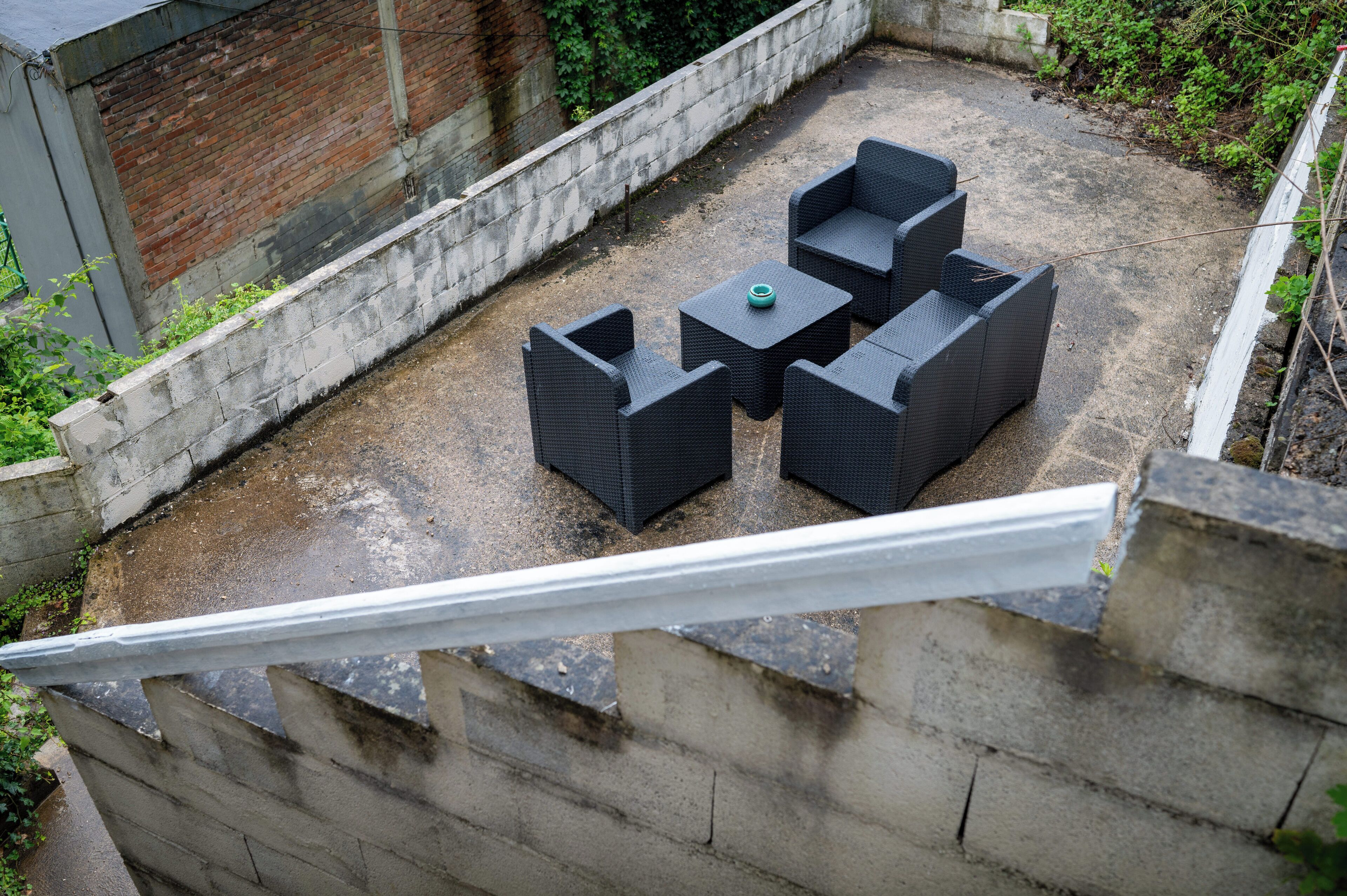 A rooftop patio with four dark wicker chairs and a small table, surrounded by a concrete wall and greenery.