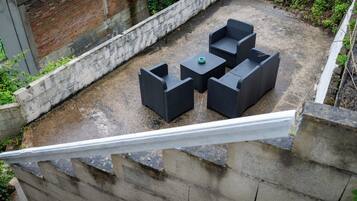 A rooftop patio with four dark wicker chairs and a small table, surrounded by a concrete wall and greenery.