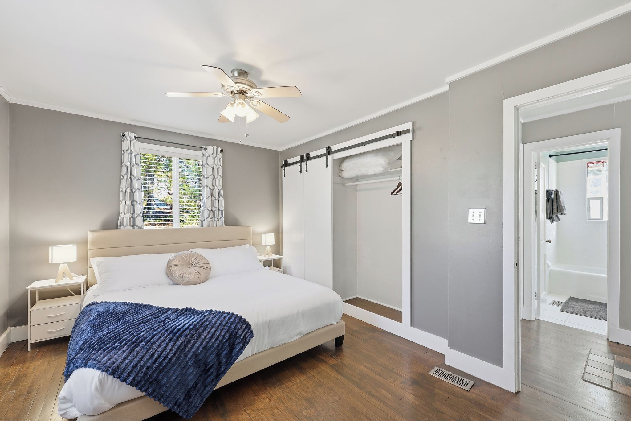A bedroom with a bed, nightstands, a ceiling fan, a window with curtains, a walk-in closet, and a bathroom door.