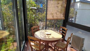 Una habitación iluminada por el sol con una mesa redonda y cuatro sillas, ventanas amplias con vistas a árboles y edificios, y un radiador en el suelo.