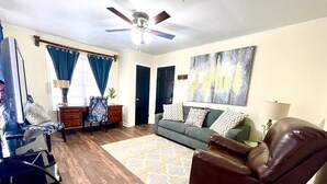 A living room with a sofa, a leather chair, a coffee table, a TV, and a ceiling fan. - Book Your HQ in LC W/Garage-FAST WiFi (Lake Charles)