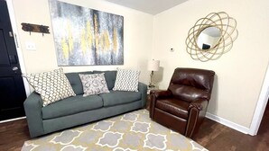 A living room with a green sofa, patterned cushions, a brown leather chair, a large abstract painting, a gold mirror, and a patterned rug. - Book Your HQ in LC W/Garage-FAST WiFi (Lake Charles)
