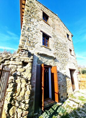 A stone building with a wooden door and window shutters, a small porch, and a stone wall.