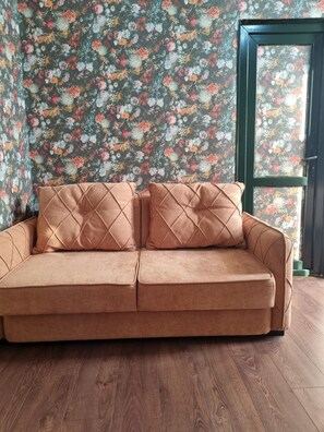 Un canapé marron orné de coussins à motifs losangés, dans une pièce dont les murs sont recouverts d’un papier peint à motifs floraux et dont le sol est en bois.