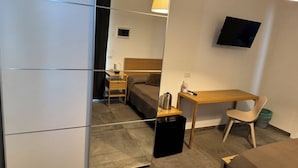 A compact hotel room with a bed, a desk, a chair, a TV mounted on the wall, and a small table with a lamp.