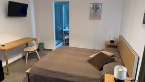 A bedroom with a bed, a desk, a chair, a TV, a bedside table with a lamp, and a picture on the wall.