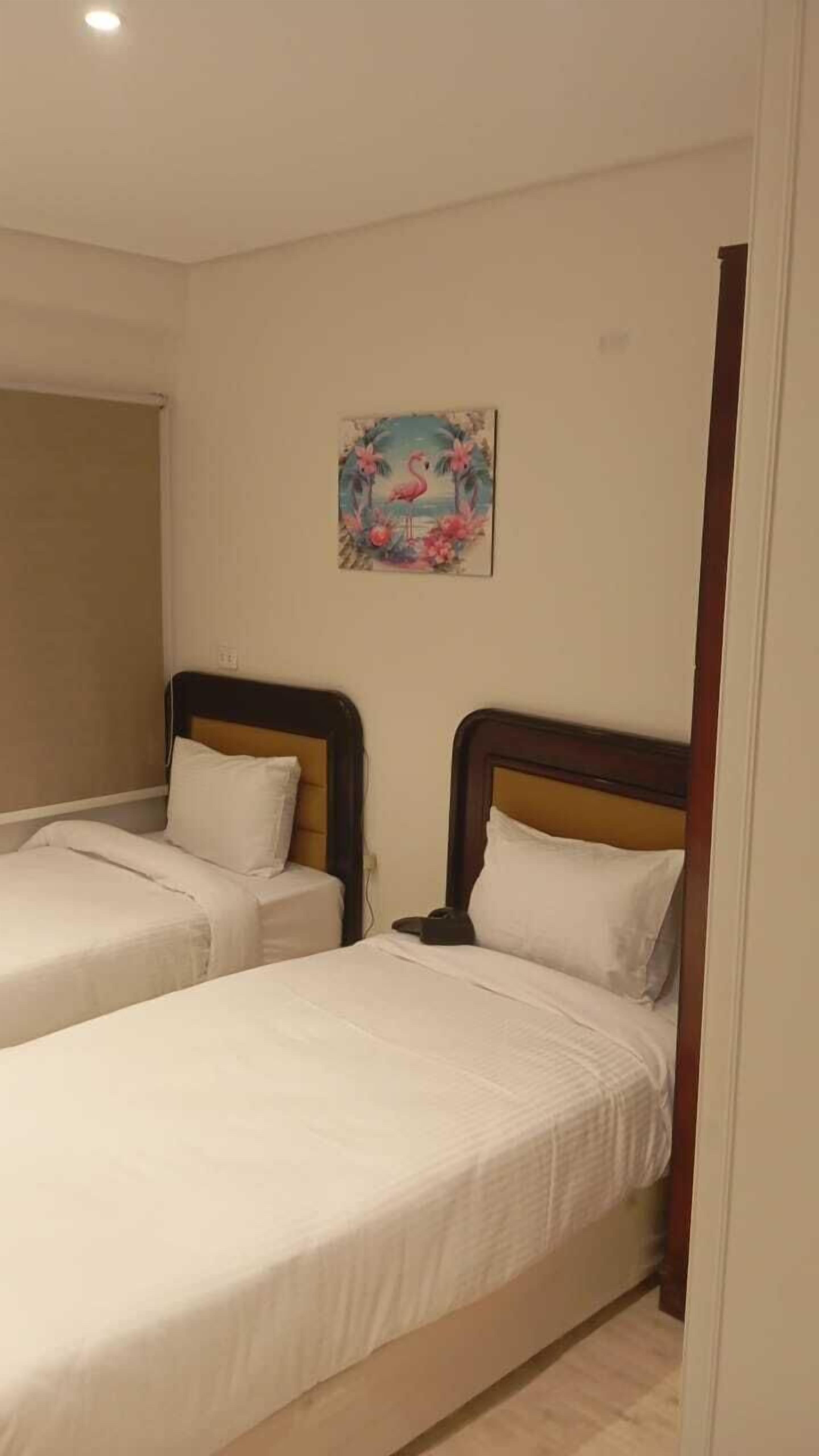 A hotel room with two single beds, a small painting on the wall, and a window with a beige blind.