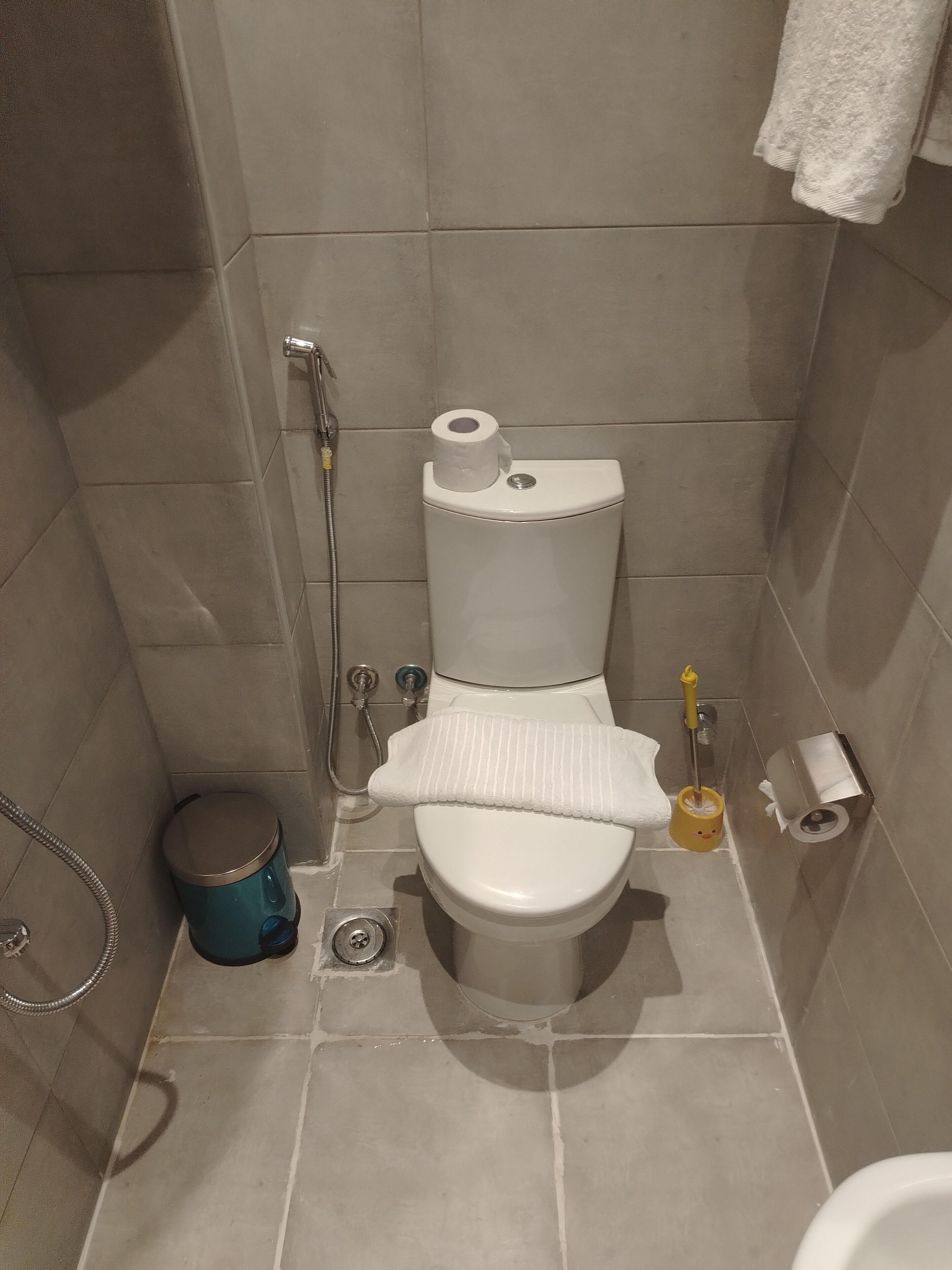 A small, tiled bathroom with a toilet, a shower, and a trash bin.
