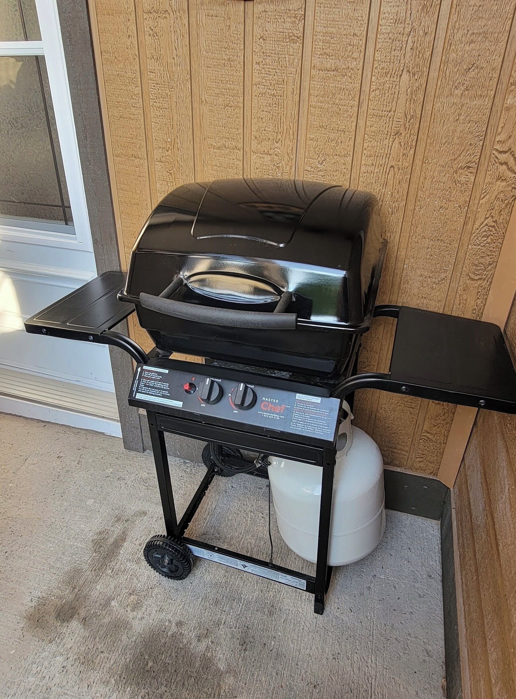 Una parrilla de gas portátil con panel de control, tanque de gas y una mesita lateral.