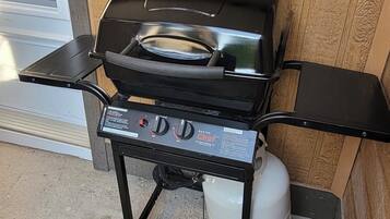 Una parrilla de gas portátil con panel de control, tanque de gas y una mesita lateral.