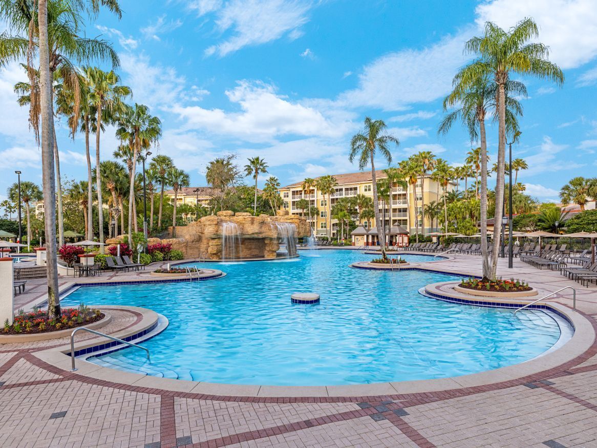 A large outdoor swimming pool with palm trees and a waterfall feature, surrounded by lounge chairs and a hotel building in the background.