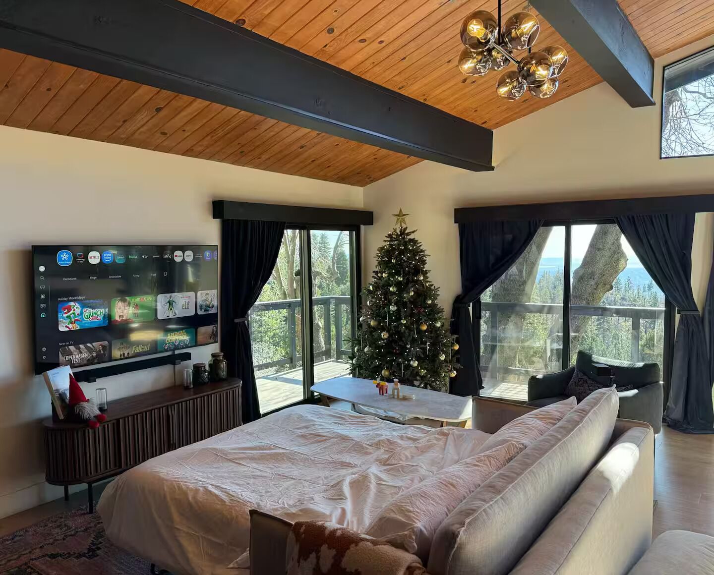 A bedroom with a large bed, a TV mounted on the wall, a Christmas tree, and a balcony with a view of trees.