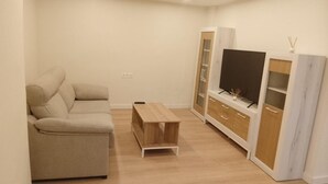 Ένα σαλόνι με έναν μπεζ καναπέ, ένα ξύλινο τραπεζάκι σαλονιού, μια τηλεόραση πάνω σε μια λευκή κονσόλα και μια λευκή ντουλάπα.