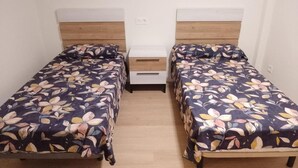 Ένα δωμάτιο με δύο μονά κρεβάτια, το καθένα με ξύλινο προσκέφαλο και ένα διακοσμητικό κάλυμμα με σχέδια λουλουδιών. Υπάρχει ένα μικρό κομοδίνο με ένα συρτάρι ανάμεσα στα κρεβάτια.