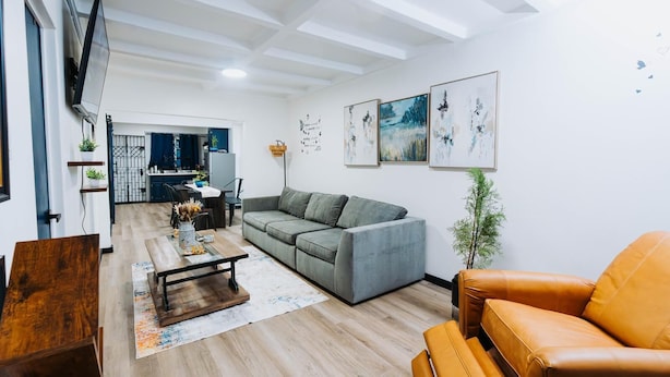 A modern living room with a grey sofa, a wooden coffee table, and a leather armchair. There is a kitchen in the background with a dining table and chairs. - Cozy Family-Oriented Townhome (Portmore)