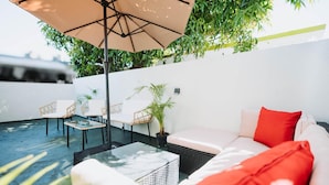 A modern outdoor seating area with a white sofa, red cushions, a glass coffee table, and a beige umbrella. - Cozy Family-Oriented Townhome (Portmore)