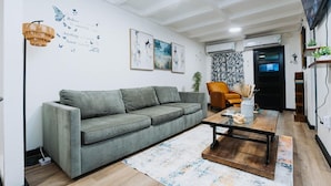 A living room with a grey sofa, a coffee table, and a floor lamp. There are air conditioning units on the wall and a door with a curtain. - Cozy Family-Oriented Townhome (Portmore)