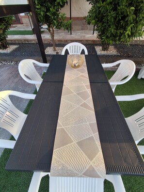 A table with a woven center runner, surrounded by white chairs, set on a patio with artificial grass and a stone border. - Casa Happy ⚠️1 to 6 guests⚠️ detached attic: 2 bedrooms + 2 bathrooms + parking  (Belpasso)