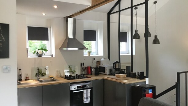 Una cucina compatta con forno, microonde e piano cottura a gas. Sono presenti finestre con tende a rullo nere, una trave in legno a soffitto e lampade a sospensione.