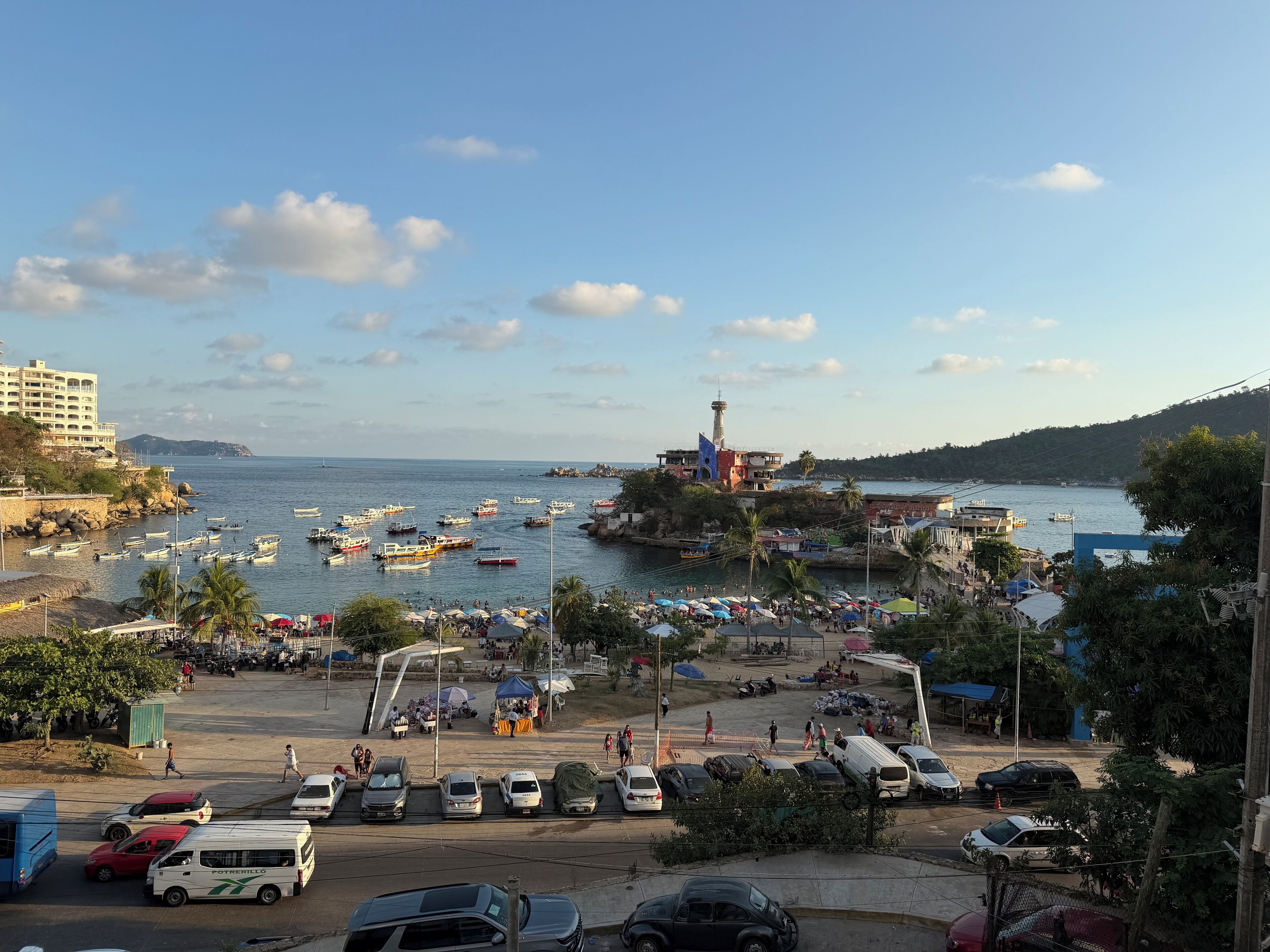 A coastal town with boats docked at the harbor, a prominent lighthouse, and a parking area with cars and a bus.
