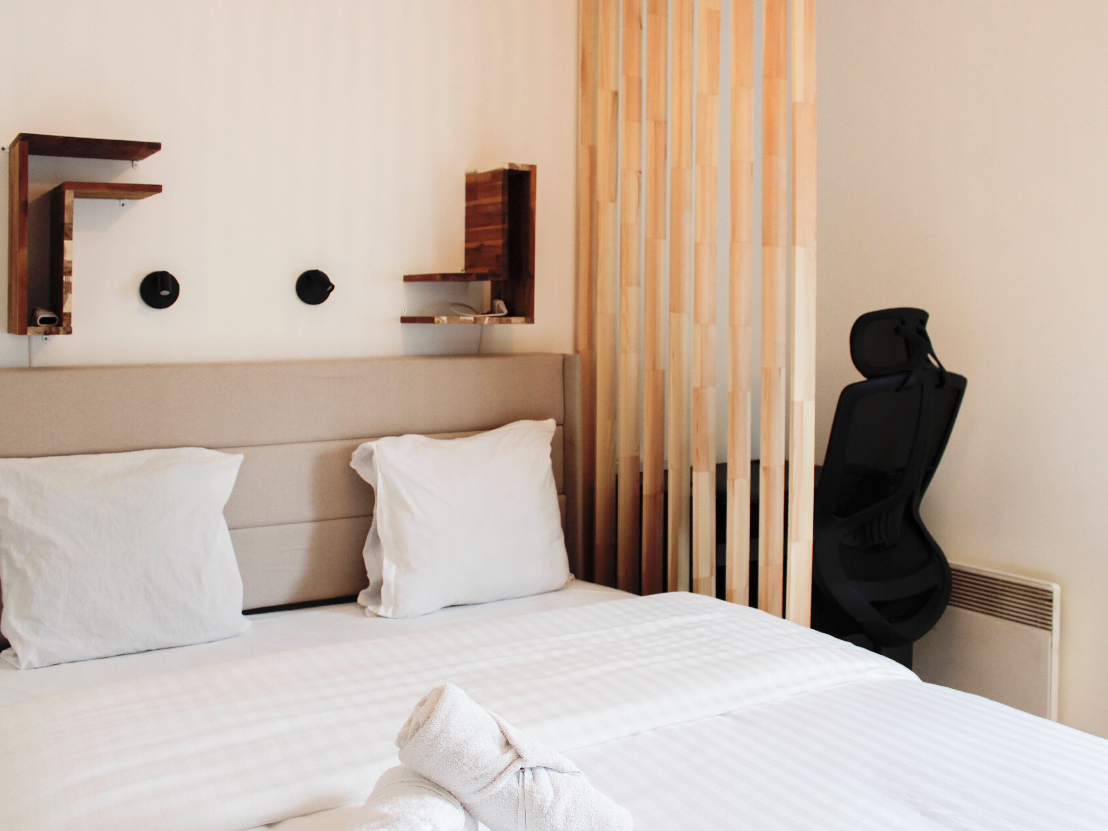 A hotel room with a bed, pillows, a towel, a chair, and a wall-mounted shelf.