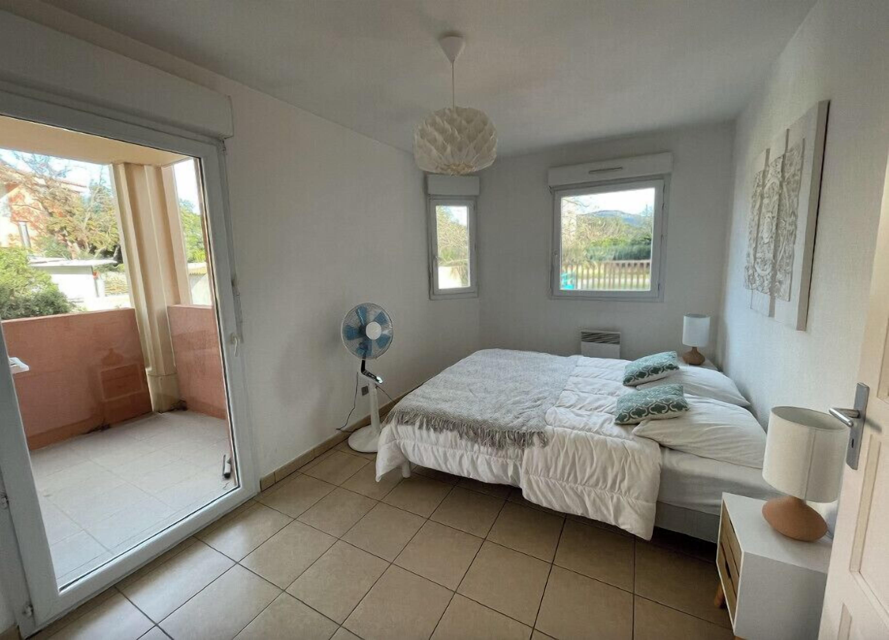 A bedroom with a bed, a standing fan, a ceiling light, and a view through a sliding door.
