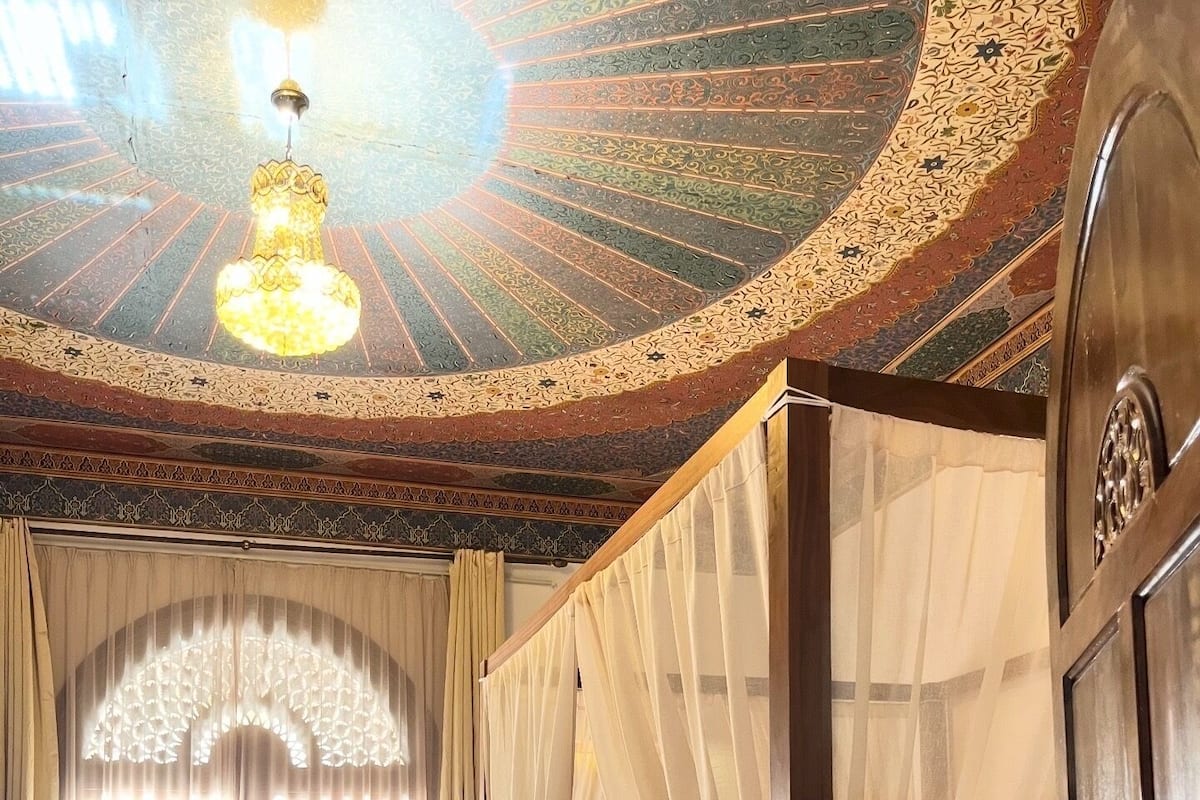 A four-poster bed with a canopy, a chandelier, and a view of a courtyard through a window with sheer curtains.