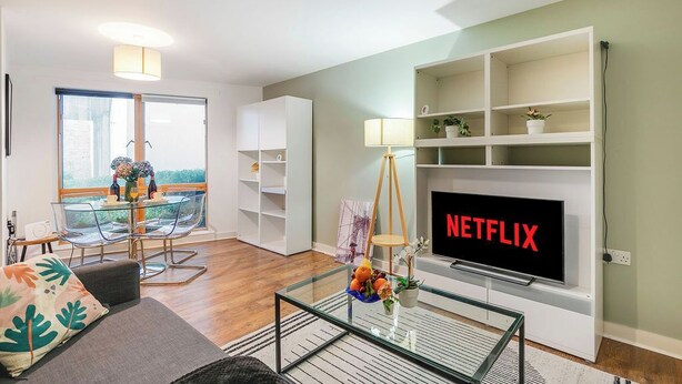 Uma sala de estar moderna com uma mesa de centro de vidro, um sofá, uma TV exibindo 'NETFLIX' e uma varanda com vista.