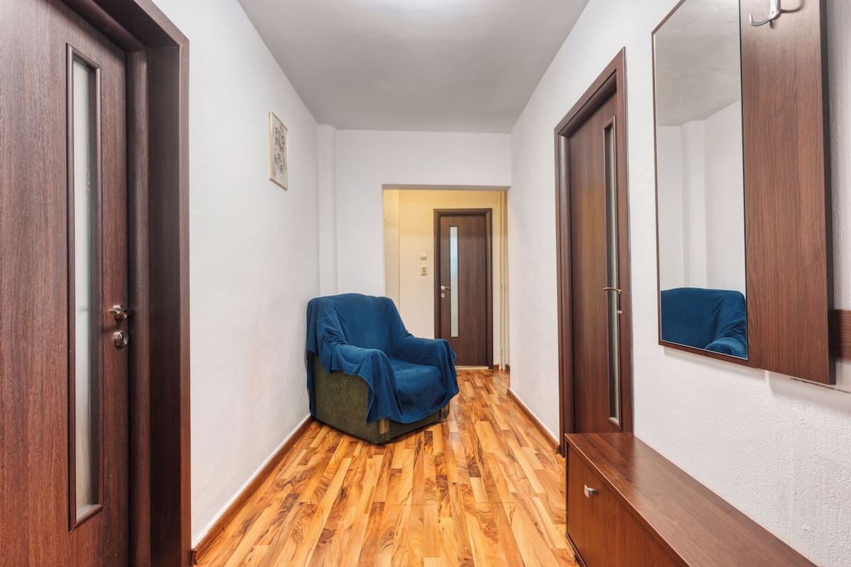 Un pasillo con suelo de madera, una silla tapizada, un espejo y una puerta.
