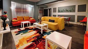 Un salon moderne avec un canapé rouge, un canapé jaune et une table basse blanche. Un tapis coloré recouvre le sol et un tableau est accroché au mur.
