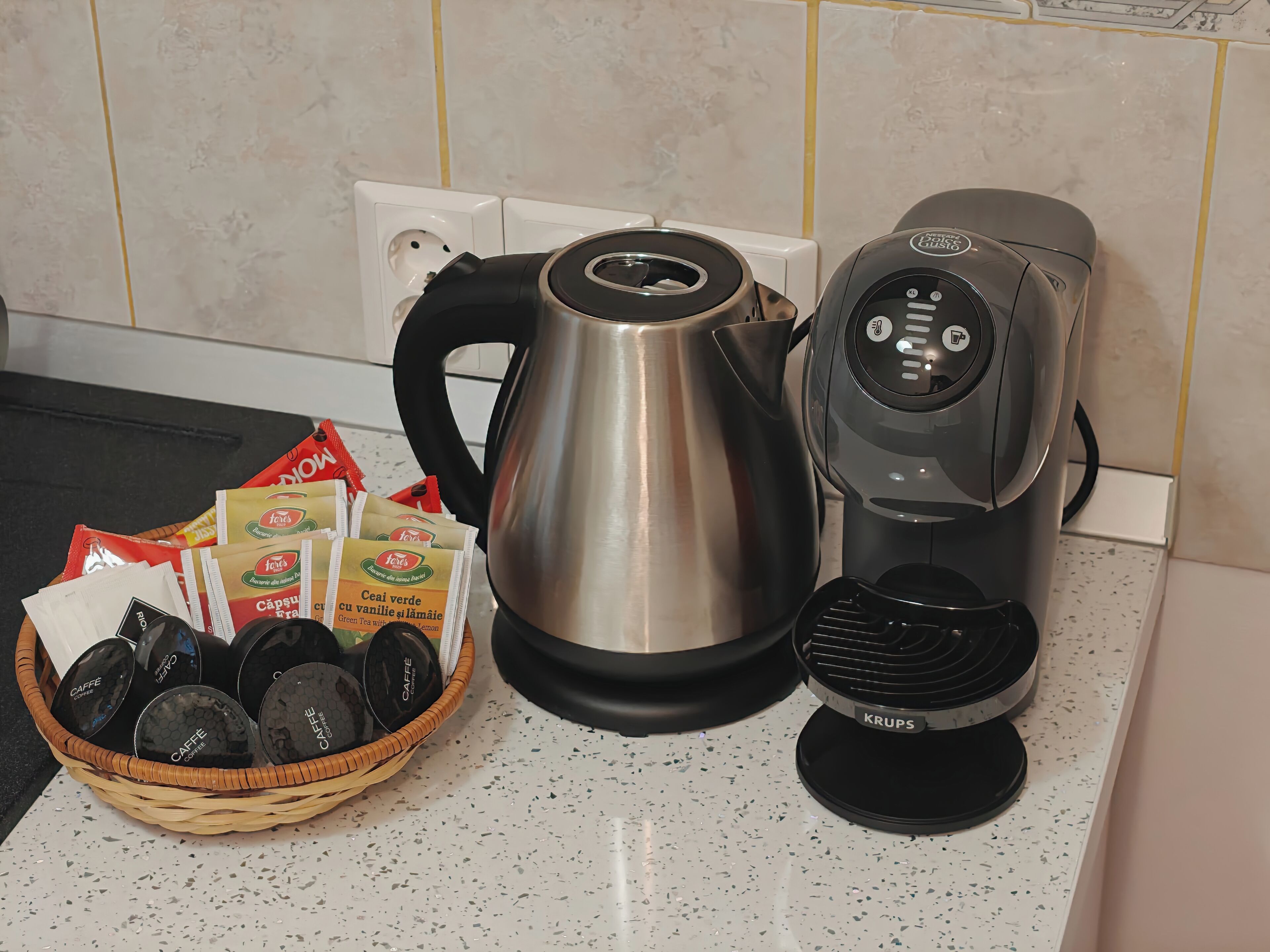 A kitchen counter with a coffee maker, a kettle, and a basket of coffee pods.