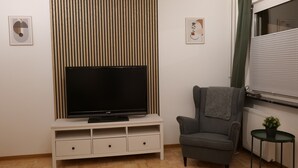 Una sala de estar con una televisión de pantalla plana sobre un mueble blanco, un sillón gris, una mesita con una planta y dos cuadros en la pared.
