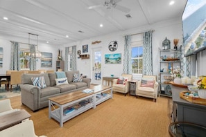 A spacious living room with a sofa, armchairs, a coffee table, and a TV. There are curtains, a bookshelf, and a window with a view.