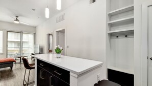 Una cocina moderna con encimera blanca, armarios negros y una pequeña isla. Hay un comedor con mesa y sillas, y un ventanal que proporciona luz natural.